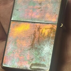 Pode incluir: Um isqueiro de metal vintage com um acabamento iridescente desgastado. A superf&iacute;cie do isqueiro exibe uma mistura de cores, incluindo laranja, verde e roxo, com arranh&otilde;es vis&iacute;veis. O design apresenta uma forma retangular com bordas arredondadas e uma tampa articulada.