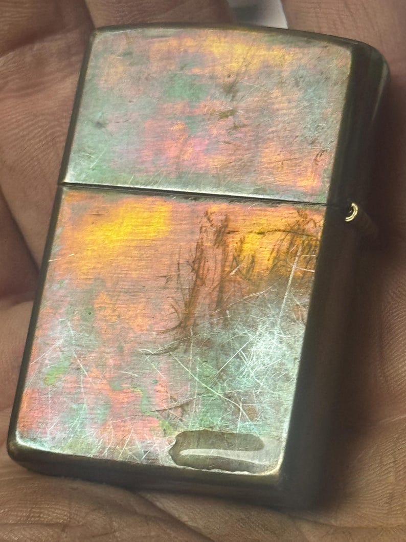 Pode incluir: Um isqueiro de metal vintage com um acabamento iridescente desgastado. A superf&iacute;cie do isqueiro exibe uma mistura de cores, incluindo laranja, verde e roxo, com arranh&otilde;es vis&iacute;veis. O design apresenta uma forma retangular com bordas arredondadas e uma tampa articulada.