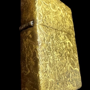 May include: A rectangular gold-colored metal lighter featuring a textured, crinkled surface pattern across its exterior. The lighter includes a prominent silver-toned hinge on the side, connecting the lid to the main body of the rectangular case.