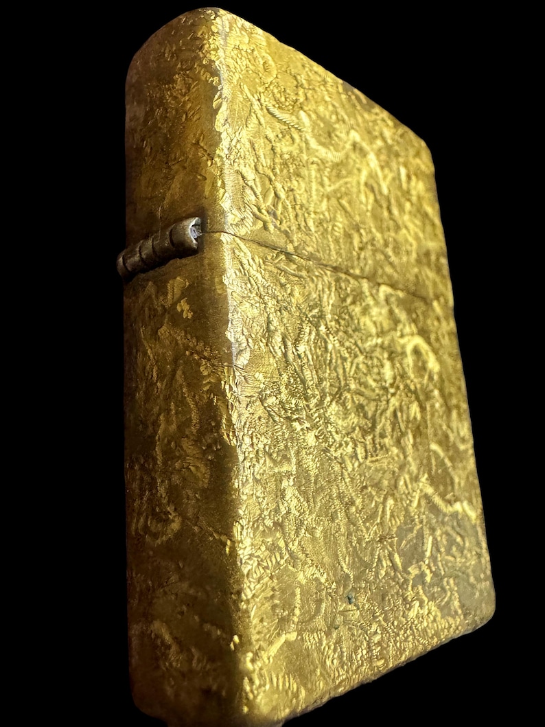 May include: A rectangular gold-colored metal lighter featuring a textured, crinkled surface pattern across its exterior. The lighter includes a prominent silver-toned hinge on the side, connecting the lid to the main body of the rectangular case.