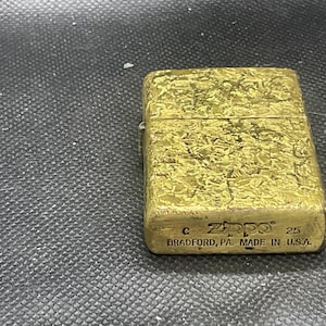 May include: A rectangular gold-toned metal lighter with a textured, weathered surface. The front features the embossed text Zippo, the letter C, the number 25, and the words BRADFORD, PA. MADE IN U.S.A. at the bottom of the lighter casing.