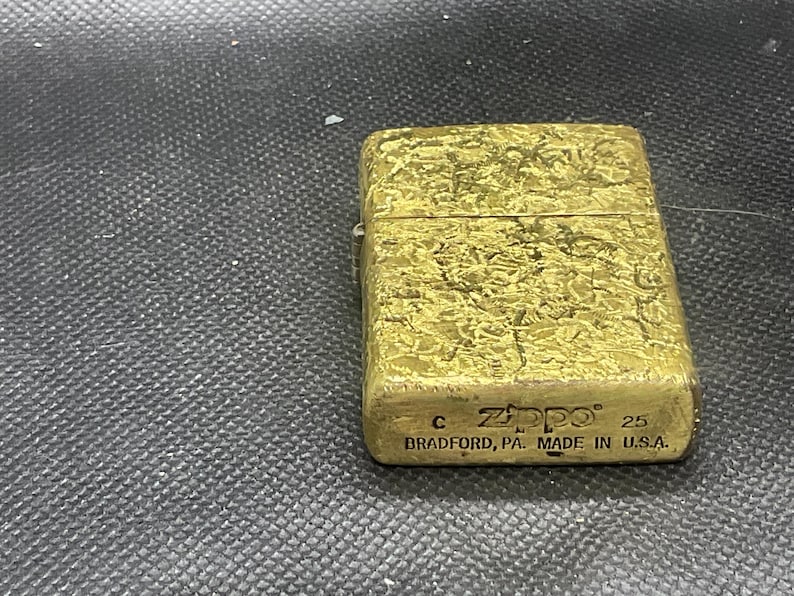 May include: A rectangular gold-toned metal lighter with a textured, weathered surface. The front features the embossed text Zippo, the letter C, the number 25, and the words BRADFORD, PA. MADE IN U.S.A. at the bottom of the lighter casing.