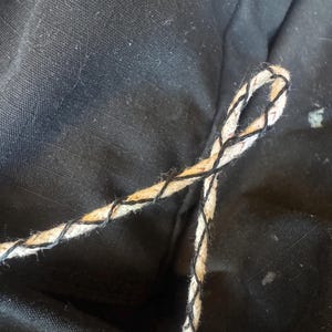 May include: Close-up of a loop of rope with a natural beige color and black stitching. The rope is against a black fabric background. The rope appears to be made of natural fibers.