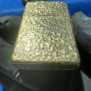 May include: A rectangular gold-toned metal lighter featuring a textured, hammered surface finish. The base of the lighter displays engraved text that reads Zippo, Bradford PA, Made in U.S.A. The object has a hinged lid and a metallic, reflective appearance.