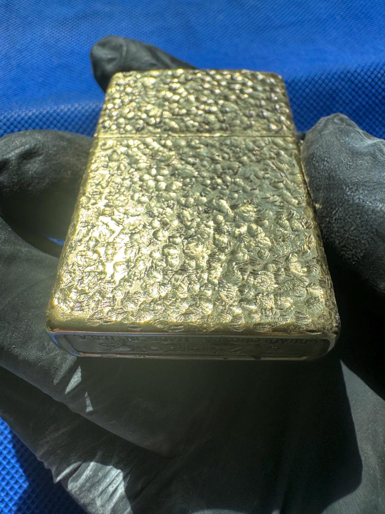 May include: A rectangular gold-toned metal lighter featuring a textured, hammered surface finish. The base of the lighter displays engraved text that reads Zippo, Bradford PA, Made in U.S.A. The object has a hinged lid and a metallic, reflective appearance.