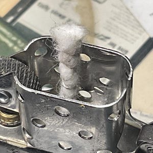 May include: A silver-toned metal lighter insert featuring a central white cotton wick surrounded by a perforated chimney. A textured circular flint wheel is positioned on the side, with a small brass-colored component visible at the base of the wheel.