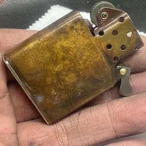 May include: A rectangular brass lighter with a weathered, mottled brown patina. The metal casing features a hinged top, a serrated ignition wheel, and a perforated chimney insert. The internal mechanism is partially visible, showing a small brass rivet.