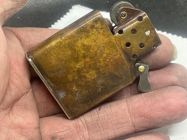 May include: A rectangular brass lighter with a weathered, mottled brown patina. The metal casing features a hinged top, a serrated ignition wheel, and a perforated chimney insert. The internal mechanism is partially visible, showing a small brass rivet.