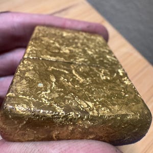 May include: A rectangular gold-colored metal lighter with a textured, uneven surface finish. The object features a hinged lid design with a horizontal seam running across the upper portion of the body, showcasing a metallic, reflective, and crinkled appearance.