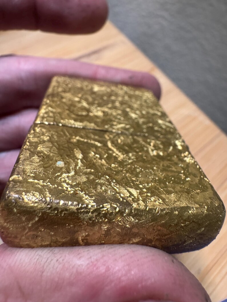 May include: A rectangular gold-colored metal lighter with a textured, uneven surface finish. The object features a hinged lid design with a horizontal seam running across the upper portion of the body, showcasing a metallic, reflective, and crinkled appearance.