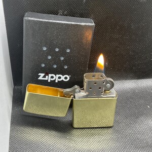 以下が含まれることがあります： 蓋が開いた真鍮製のZippoライター。炎と金属製のチムニーが見える。ライターの後ろには、テクスチャー加工された表面と小さな円形のくぼみがある、黒い長方形のZippoブランドの箱が置かれている。