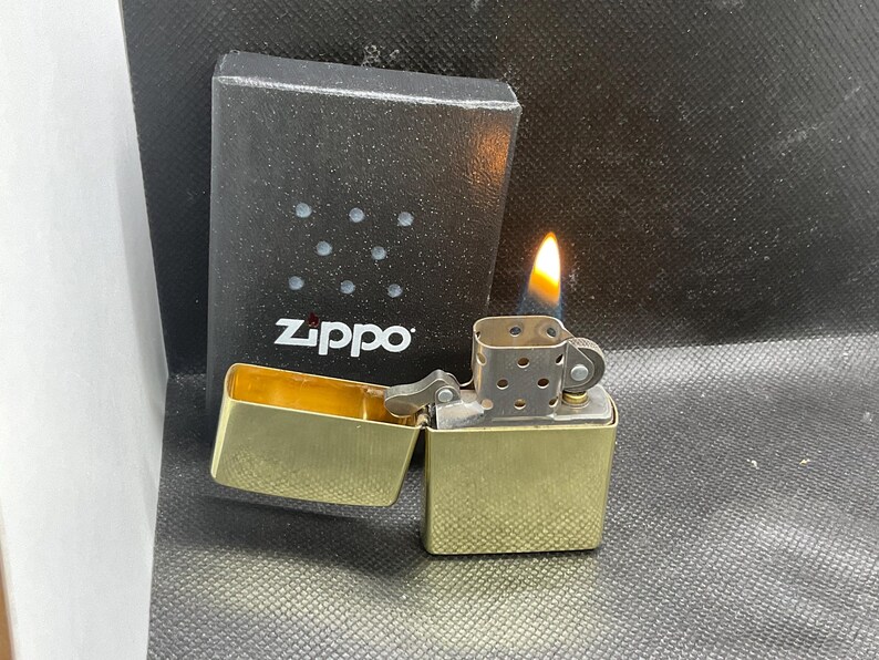 以下が含まれることがあります： 蓋が開いた真鍮製のZippoライター。炎と金属製のチムニーが見える。ライターの後ろには、テクスチャー加工された表面と小さな円形のくぼみがある、黒い長方形のZippoブランドの箱が置かれている。