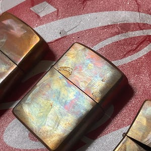 Pode incluir: Close-up de tr&ecirc;s isqueiros de metal vintage com um acabamento iridescente e desgastado. Os isqueiros t&ecirc;m formato retangular com bordas arredondadas e uma tampa articulada. As cores variam do bronze ao dourado, com toques de azul e rosa.