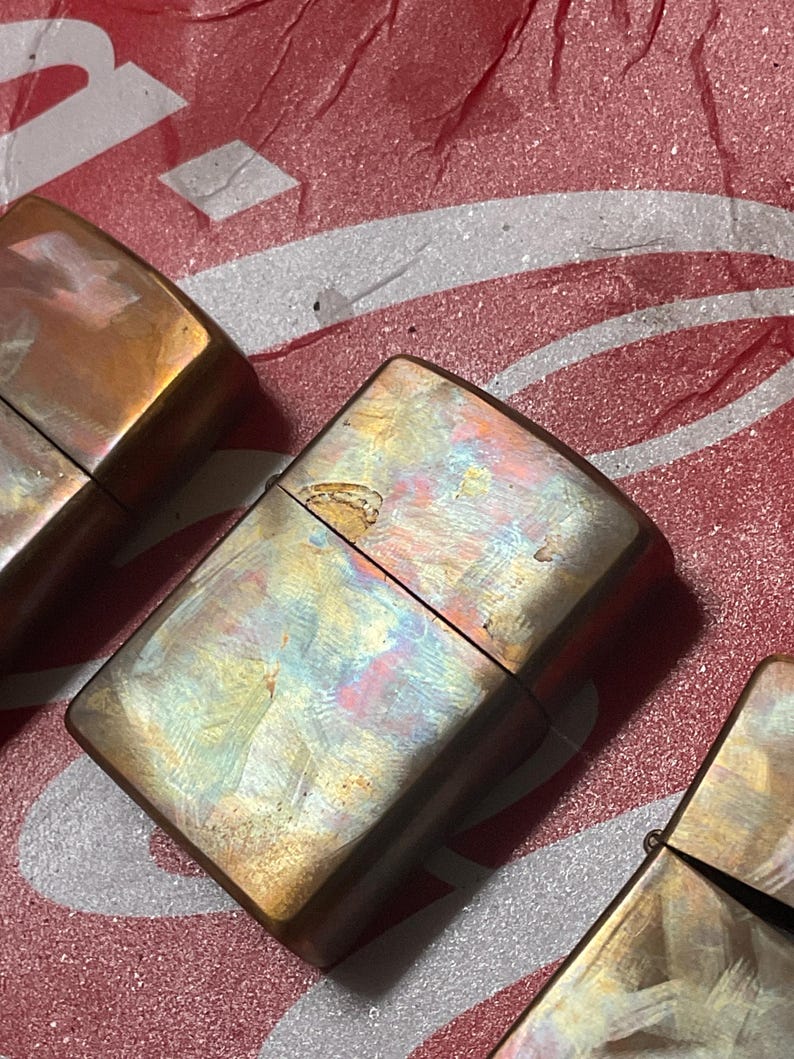Pode incluir: Close-up de tr&ecirc;s isqueiros de metal vintage com um acabamento iridescente e desgastado. Os isqueiros t&ecirc;m formato retangular com bordas arredondadas e uma tampa articulada. As cores variam do bronze ao dourado, com toques de azul e rosa.