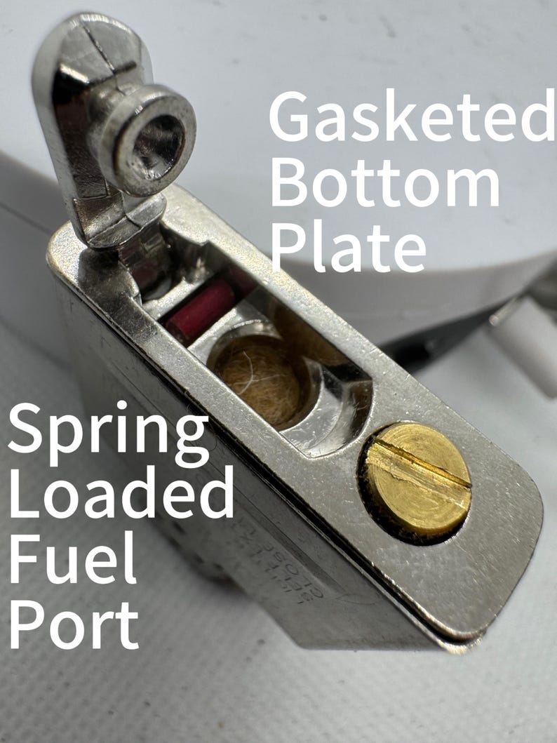 May include: A metallic lighter insert featuring a Gasketed Bottom Plate and a Spring Loaded Fuel Port. The component includes a brass-colored screw, a small red rectangular element, and a central circular opening containing fibrous material.