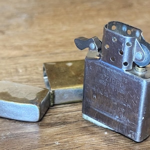 May include: A vintage silver Zippo lighter with an open lid, revealing the internal components. The lighter's case is slightly worn, with visible text and a brass-colored insert. The lid is detached, lying next to the lighter.