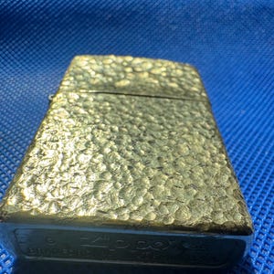 May include: A gold-toned metal lighter with a textured, hammered surface. The bottom of the lighter features the engraved text Zippo, Bradford PA, MADE IN U.S.A., along with a G and 25 stamp, indicating its manufacturing details and origin.