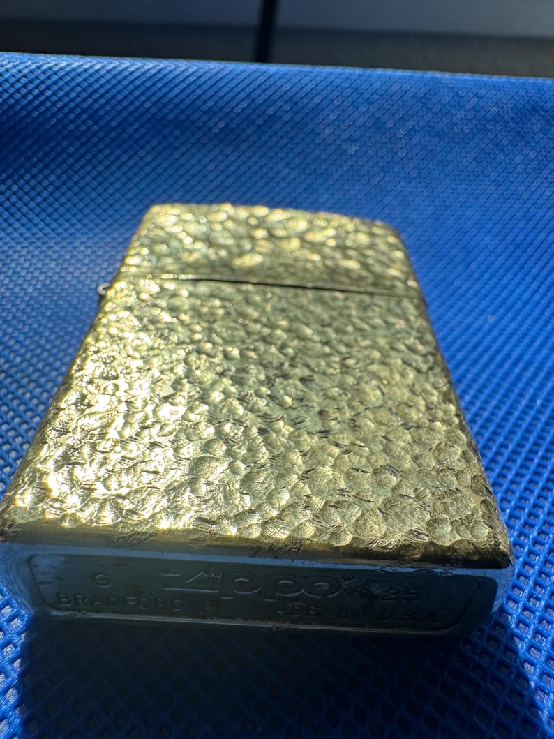 May include: A gold-toned metal lighter with a textured, hammered surface. The bottom of the lighter features the engraved text Zippo, Bradford PA, MADE IN U.S.A., along with a G and 25 stamp, indicating its manufacturing details and origin.