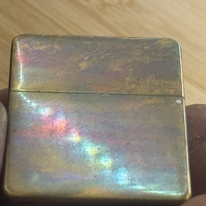 Pode incluir: Um isqueiro retangular vintage com acabamento met&aacute;lico. A superf&iacute;cie do isqueiro tem uma apar&ecirc;ncia desgastada com reflexos iridescentes coloridos do arco-&iacute;ris. O isqueiro &eacute; segurado em uma m&atilde;o, e o fundo &eacute; uma superf&iacute;cie de madeira.