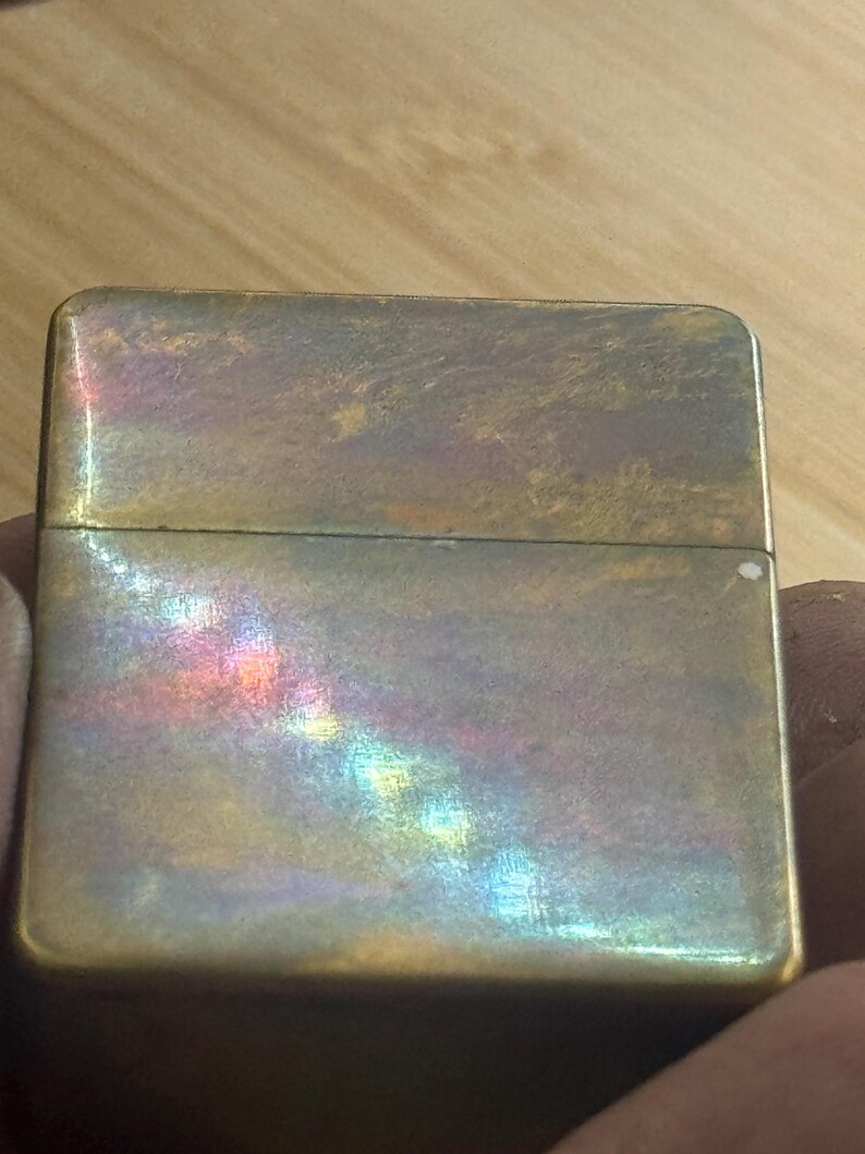 Pode incluir: Um isqueiro retangular vintage com acabamento met&aacute;lico. A superf&iacute;cie do isqueiro tem uma apar&ecirc;ncia desgastada com reflexos iridescentes coloridos do arco-&iacute;ris. O isqueiro &eacute; segurado em uma m&atilde;o, e o fundo &eacute; uma superf&iacute;cie de madeira.