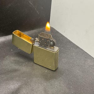 以下が含まれることがあります： 蓋が開いた状態のブラッシュドブラス製ライター。銀色のチムニーインサートと、芯から揺らめく小さな明るいオレンジと黄色の炎が特徴です。長方形のライターは、クラシックなフリップトップデザインと金属仕上げが施されています。