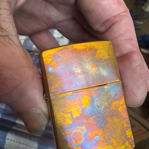 Pode incluir: Um isqueiro met&aacute;lico retangular com um acabamento iridescente. A superf&iacute;cie exibe uma mistura de tons dourados, laranja, azul e roxo, criando uma apar&ecirc;ncia texturizada &uacute;nica. O isqueiro &eacute; segurado em uma m&atilde;o, mostrando seu tamanho compacto e design.