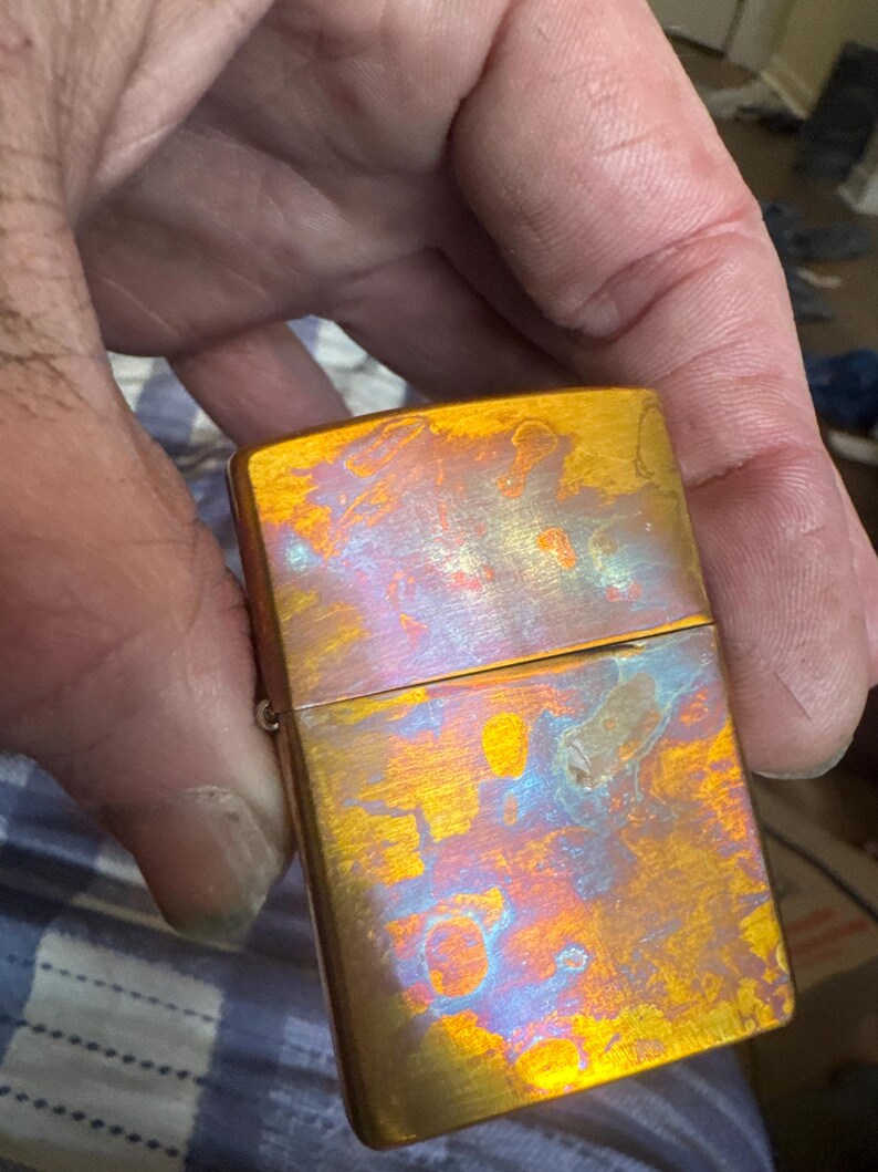 Pode incluir: Um isqueiro met&aacute;lico retangular com um acabamento iridescente. A superf&iacute;cie exibe uma mistura de tons dourados, laranja, azul e roxo, criando uma apar&ecirc;ncia texturizada &uacute;nica. O isqueiro &eacute; segurado em uma m&atilde;o, mostrando seu tamanho compacto e design.