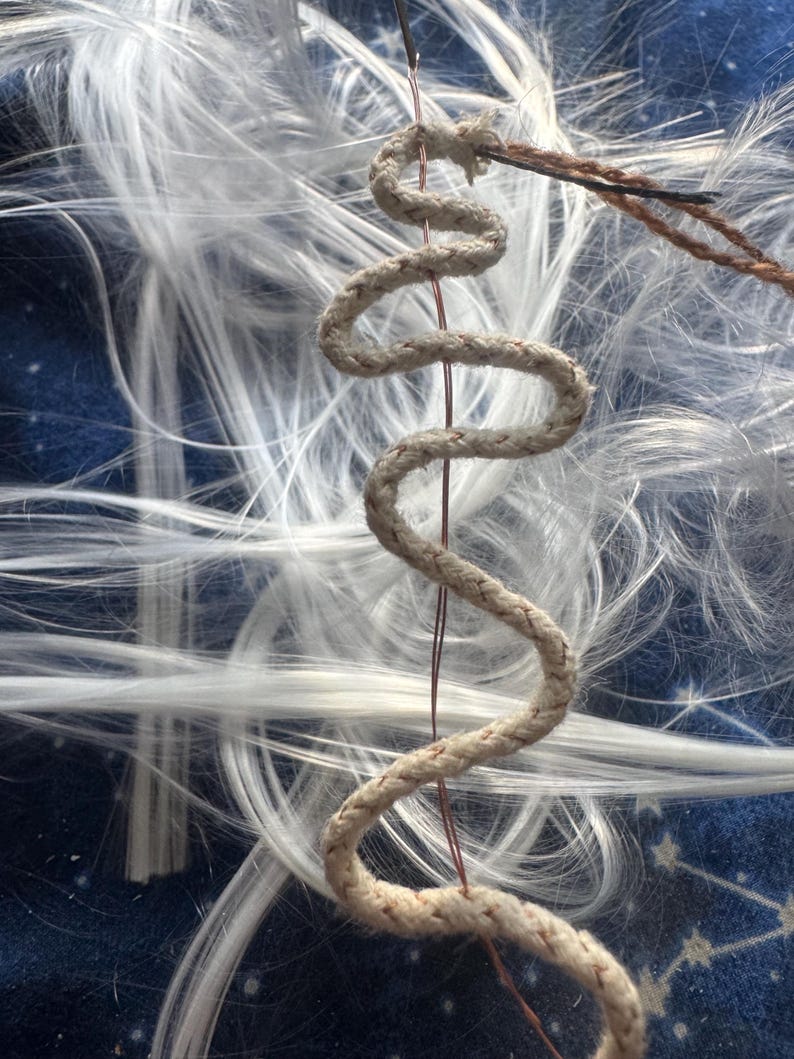 May include: A beige, braided rope shaped into a vertical, serpentine wave pattern. A thin, dark copper-colored wire runs through the center of the rope, extending from the top and bottom. The rope is positioned against a background of white fibers.