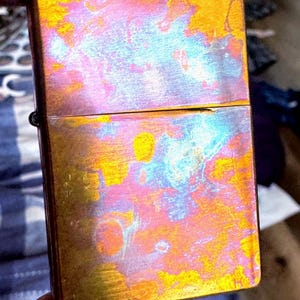 Pode incluir: Um isqueiro retangular iridescente com tampa articulada. A superf&iacute;cie do isqueiro exibe uma mistura vibrante de cores, incluindo tons de ouro, rosa e azul. O isqueiro &eacute; segurado na m&atilde;o de uma pessoa.