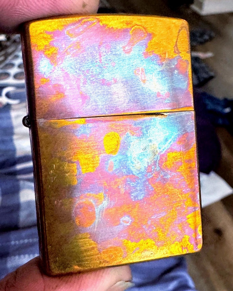 Pode incluir: Um isqueiro retangular iridescente com tampa articulada. A superf&iacute;cie do isqueiro exibe uma mistura vibrante de cores, incluindo tons de ouro, rosa e azul. O isqueiro &eacute; segurado na m&atilde;o de uma pessoa.