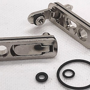 May include: Two rectangular silver-toned metal compression latches with a polished finish, each featuring a central lever mechanism and a circular mounting hole. Included are three black rubber O-rings of varying sizes for installation and sealing.