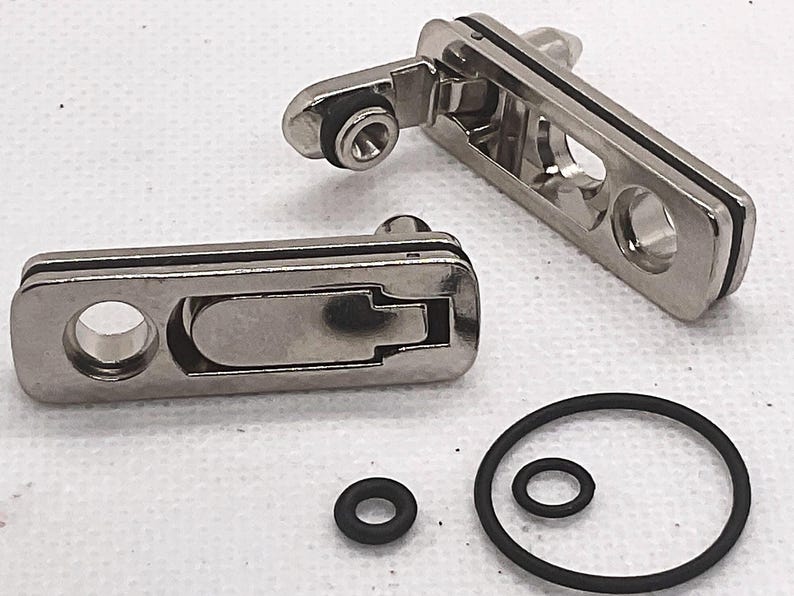 May include: Two rectangular silver-toned metal compression latches with a polished finish, each featuring a central lever mechanism and a circular mounting hole. Included are three black rubber O-rings of varying sizes for installation and sealing.