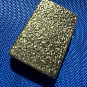 May include: A rectangular metal lighter with a gold-toned finish and a distinct hammered texture across its entire surface. The lighter features a horizontal hinge line separating the lid from the main body, creating a classic flip-top design.