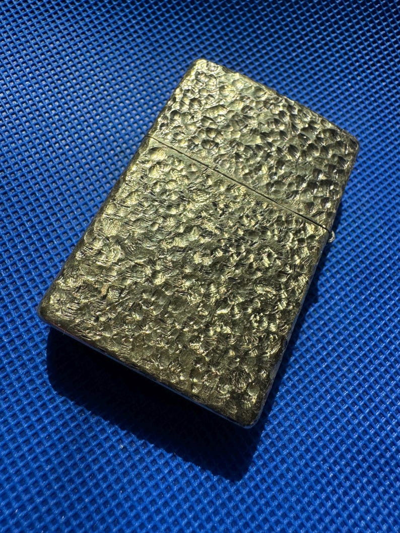 May include: A rectangular metal lighter with a gold-toned finish and a distinct hammered texture across its entire surface. The lighter features a horizontal hinge line separating the lid from the main body, creating a classic flip-top design.
