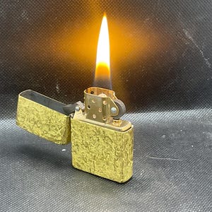 May include: A gold-toned metal flip-top lighter with a textured, hammered surface finish. The lighter is open with its lid tilted back, and a bright orange and yellow flame is actively burning from the wick, illuminating the metallic casing.