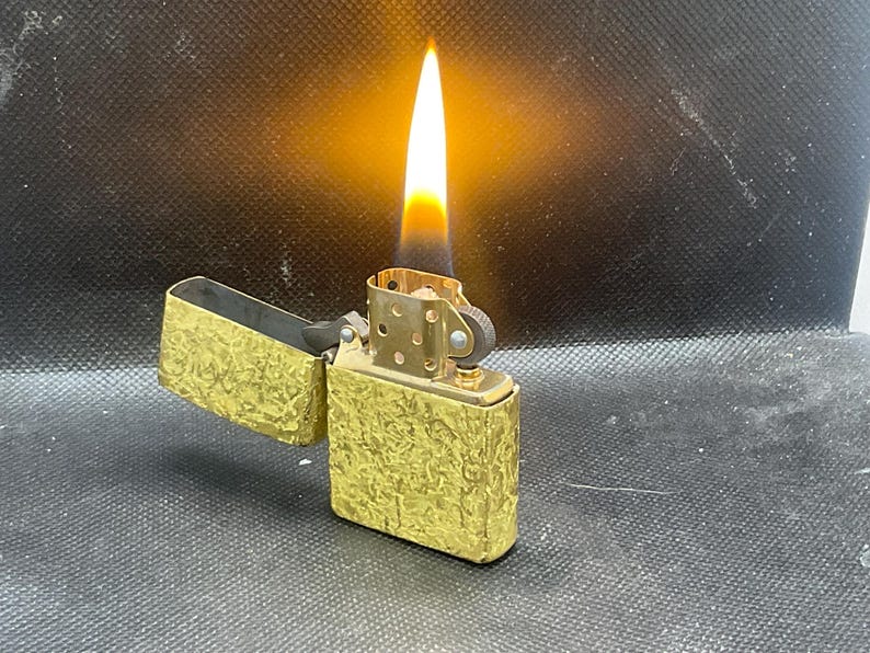 May include: A gold-toned metal flip-top lighter with a textured, hammered surface finish. The lighter is open with its lid tilted back, and a bright orange and yellow flame is actively burning from the wick, illuminating the metallic casing.