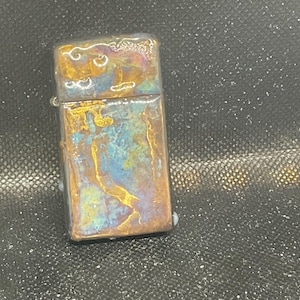 Pode incluir: Um isqueiro retangular com acabamento met&aacute;lico. O isqueiro tem um padr&atilde;o de cores &uacute;nico com tons de ouro, azul e roxo. A parte superior do isqueiro tem uma apar&ecirc;ncia texturizada.