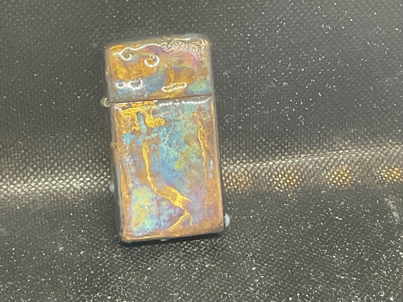 Pode incluir: Um isqueiro retangular com acabamento met&aacute;lico. O isqueiro tem um padr&atilde;o de cores &uacute;nico com tons de ouro, azul e roxo. A parte superior do isqueiro tem uma apar&ecirc;ncia texturizada.