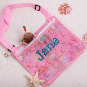 May include: Pink mesh beach bag with a floral pattern and the name "Jane" embroidered in teal. The bag has a pink strap and a zipper closure. Sunglasses and seashells are also visible.
