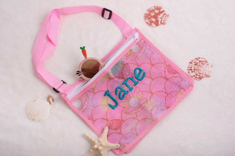 May include: Pink mesh beach bag with a floral pattern and the name "Jane" embroidered in teal. The bag has a pink strap and a zipper closure. Sunglasses and seashells are also visible.