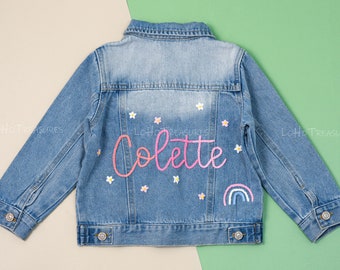 Könnte beinhalten: Eine hellblaue Jeansjacke mit dem Namen "Colette", der in rosa und weißem Garn auf der Rückseite gestickt ist. Die Jacke ist mit kleinen weißen Blumen und einem Regenbogen verziert.