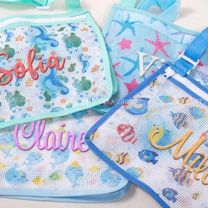 Op de afbeelding: Mesh strandtassen met gepersonaliseerde namen. De tassen hebben aquatische patronen met zeepaardjes, vissen en zeesterren. Elke tas heeft een andere naam in een sierlijk lettertype: Sofia, Claire, Mila en V. De tassen hebben blauwe en aquatinten.