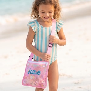 Peut inclure: Un maillot de bain à rayures bleu clair et blanc avec des manches à volants, accompagné d'un sac bandoulière en maille rose doté d'une fermeture éclair blanche et du prénom Jane brodé en écriture turquoise sur le devant du sac.