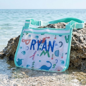 May include: A turquoise mesh beach bag with the name "RYAN" in blue letters. The bag has a white zip and an adjustable strap. It is decorated with colourful dinosaur and plant illustrations.