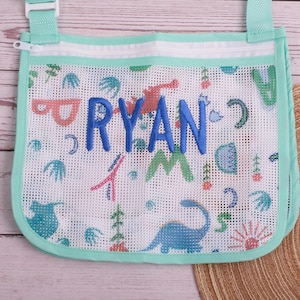 May include: A white mesh bag with a mint green trim and strap. The bag features a colorful dinosaur pattern and the name "RYAN" embroidered in blue. The bag has a zipper closure and is designed for carrying items.