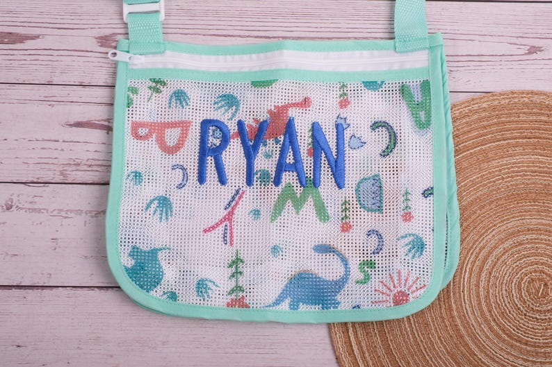 May include: A white mesh bag with a mint green trim and strap. The bag features a colorful dinosaur pattern and the name "RYAN" embroidered in blue. The bag has a zipper closure and is designed for carrying items.