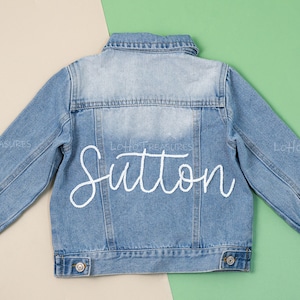 May include: A light blue denim jacket with the name "Sutton" embroidered on the back in white.