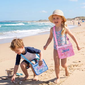 Peut inclure: Deux sacs bandoulière rectangulaires en plastique transparent avec des sangles réglables. Un sac présente un motif d'écailles rose avec le prénom Jane en bleu. L'autre sac présente un motif nautique bleu avec le prénom Leila en rose.