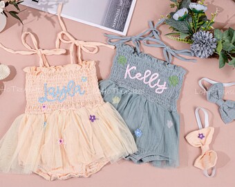 May include: Two baby rompers, one peach and one teal, with matching headbands and a bunny rattle. The peach romper has the name "Layla" embroidered in blue, and the teal romper has "Kelly" embroidered in pink.