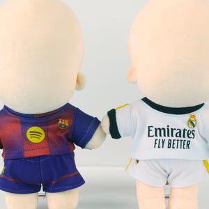 Peut inclure: Deux poupées en peluche vêtues d'uniformes de football. L'une porte un maillot bleu et rouge avec un logo Spotify jaune. L'autre porte un maillot blanc avec le texte "Emirates FLY BETTER". Les deux poupées ont la peau claire.